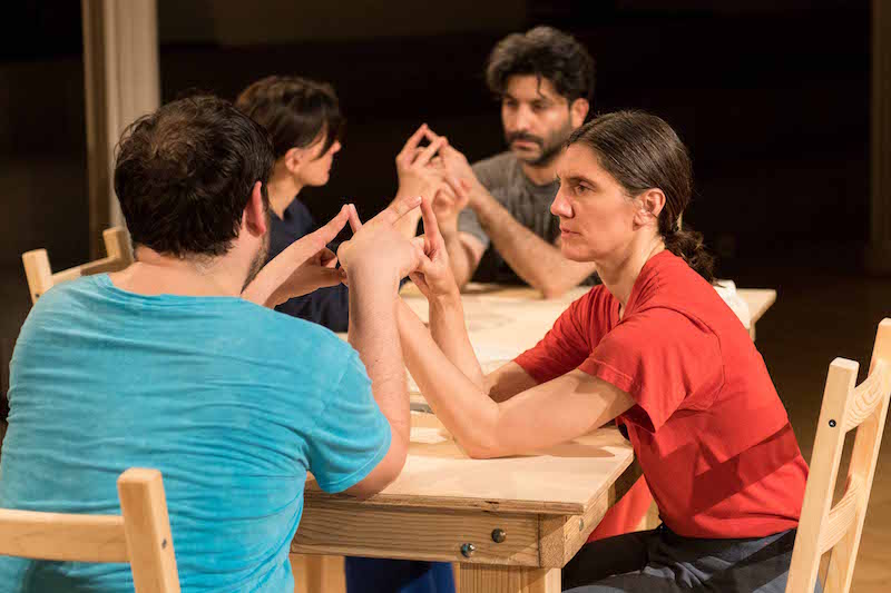 The four dancers sit around the wooden table. They pair off connecting fingers together.